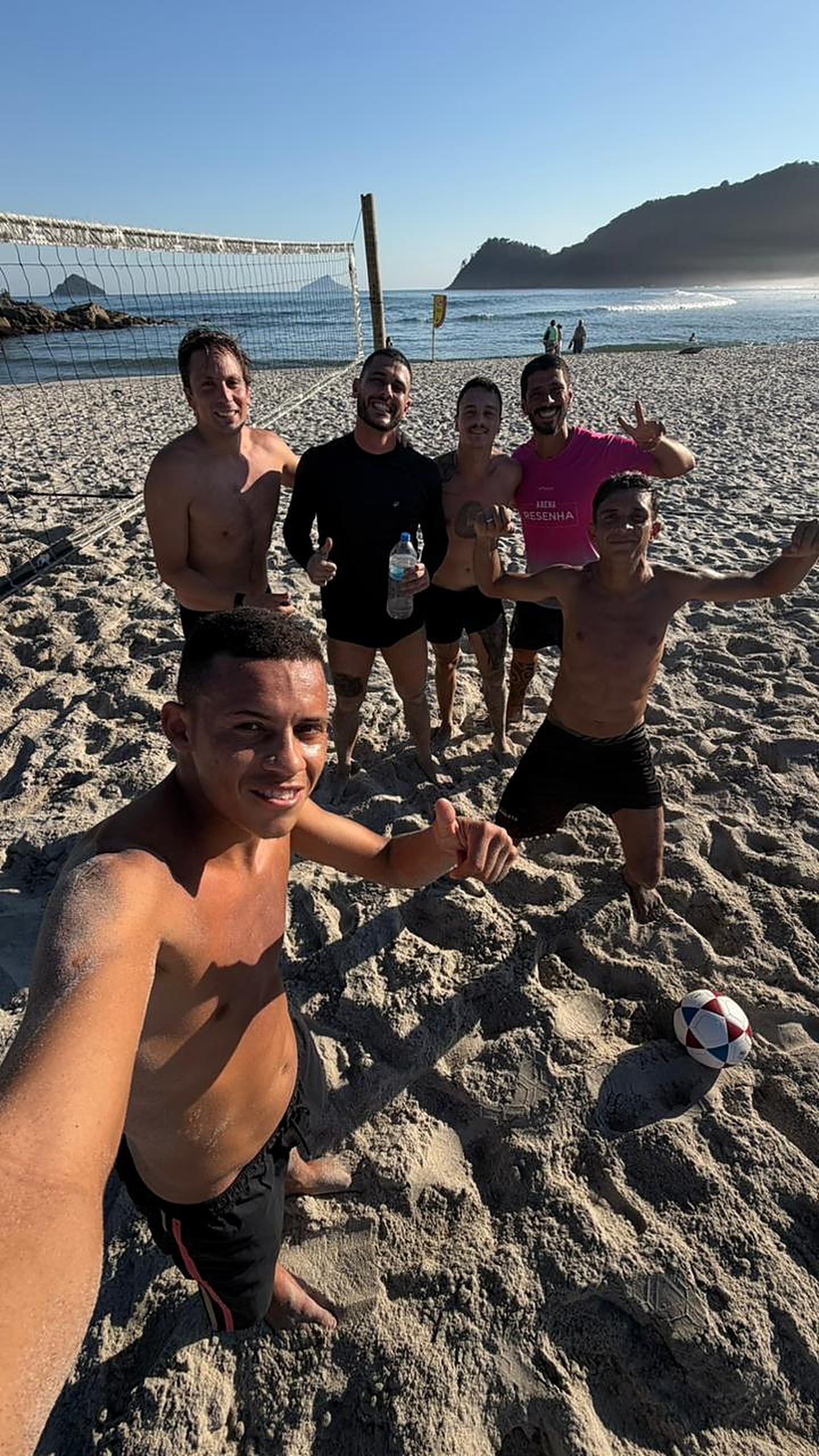Grupo de alunos do CT Dudu Futevôlei comemorando na areia com o mar ao fundo